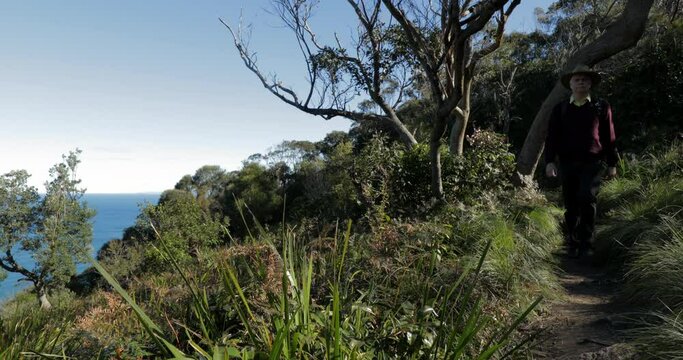 Man in hat walking on cliff-top path, with ocean below to the left hand side.  Locked down. Back pack and blimp. Bush walk in nature. Exercise in the great outdoors. Healthy lifestyle.