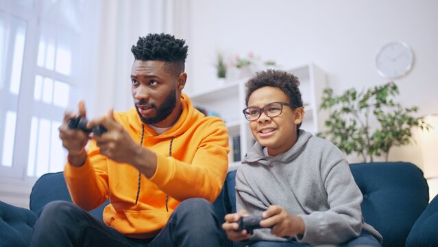 Friendly African American brothers playing videogames together at home, siblings having fun