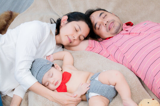 Happy Asian father and mother resting with a newborn baby boy on the bed in the bedroom. Parents and little son spend time together on a family day. Family relationship concept.