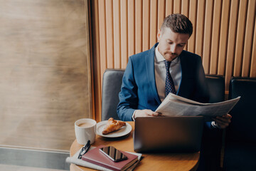 Focused entrepreneur in formal wear reads financial chronicle, coffee break with cappuccino, croissant, laptop open.