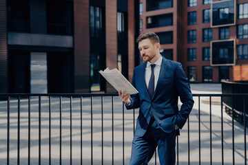 Handsome boss in blue suit stands thoughtfully, hand in pocket, reading newspaper for firm-related news, corporate buildings behind.