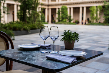 foreground on elegant dark marble table set for two, two empty crystal goblets, a seedling; in the blurred background a courtyard with colonnade and plants