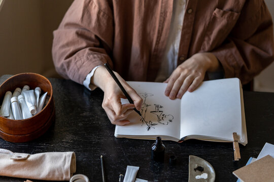 Unrecognizable female artist drawing with ink on white paper, sitting at table with paints, brushes and various art supplies, creating modern artwork in cozy art studio. Talent and creativity - Powered by Adobe