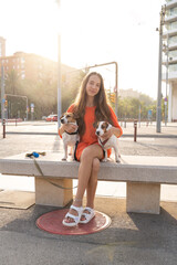 Adorable teenager girl sitting on the bench with her two Jack Russell terriers dogs. Bright orange color casual sporty clothes. Summer evening time walking with dogs in big city. 