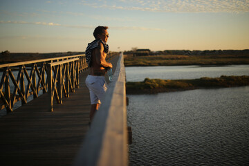 Um momento de contempla&ccedil;&atilde;o: um belo homem bronzeado aprecia o p&ocirc;r do sol sobre o passadi&ccedil;o da Ria Formosa, no Algarve, durante o ver&atilde;o em Portugal, desfrutando da tranquilidade e da beleza da paisagem