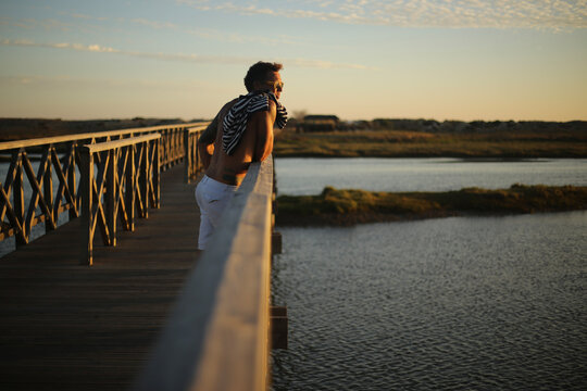 Um momento de contempla&ccedil;&atilde;o: um belo homem bronzeado aprecia o p&ocirc;r do sol sobre o passadi&ccedil;o da Ria Formosa, no Algarve, durante o ver&atilde;o em Portugal, desfrutando da tranquilidade e da beleza da paisagem