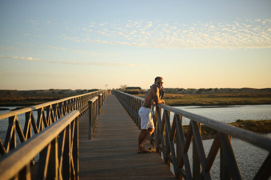 Um momento de contempla&ccedil;&atilde;o: um belo homem bronzeado aprecia o p&ocirc;r do sol sobre o passadi&ccedil;o da Ria Formosa, no Algarve, durante o ver&atilde;o em Portugal, desfrutando da tranquilidade e da beleza da paisagem