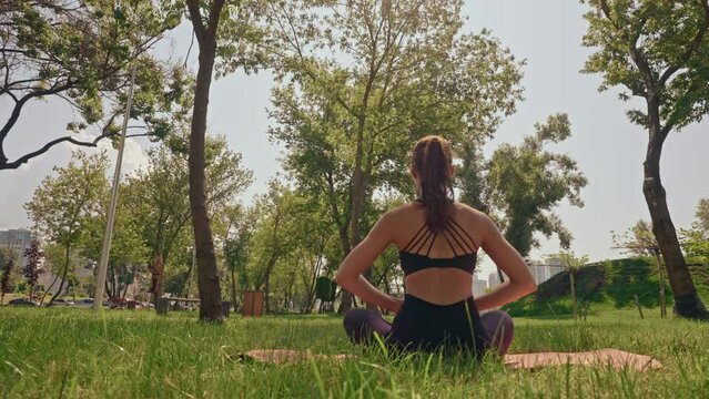 rear view woman sitting on yoga mat in lotus pose breathing exercises in park summer sunny day