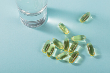 Diet, nutrition, healthy eating concept. Oil filled capsules of food supplements and glass of water for pouring yellow softgels capsules on light blue background.