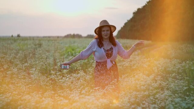 Caucasian woman in a hat dancing in a field at sunset. Concept of smiles and happy free girl who feels freedom moving in a dance on the grass in nature. High quality 4k footage