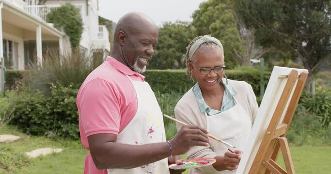 Happy senior african american couple painting on wooden easel in garden, slow motion