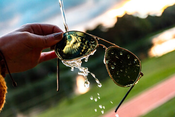  conceptual image of water falling on dark sunglasses at sunset. woman's hands holding aviator glasses. woman holding black sunglasses with water spraying on them. water in motion.