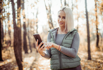 Smiling fitness blonde woman using smartphone in autumn forest