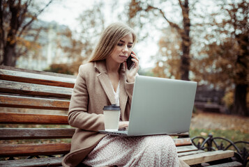 Young business woman in coat works with a laptop,  talking on phone while sitting on bench in city...