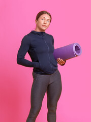 Athletic woman trainer in sportswear with a mat in her hands on a pink studio background. Healthy lifestyle. Sport, bodybuilding and fitness concept