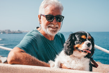 Portrait of smiling white haired senior man sitting close to the sea with his cavalier king charles spaniel dog. Elderly bearded man  and his best friend enjoying good time