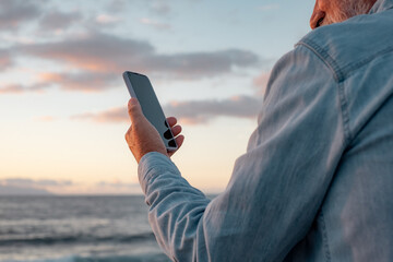 Rear view of mature caucasian man face to the sea holding mobile phone in one hand. Horizon over...