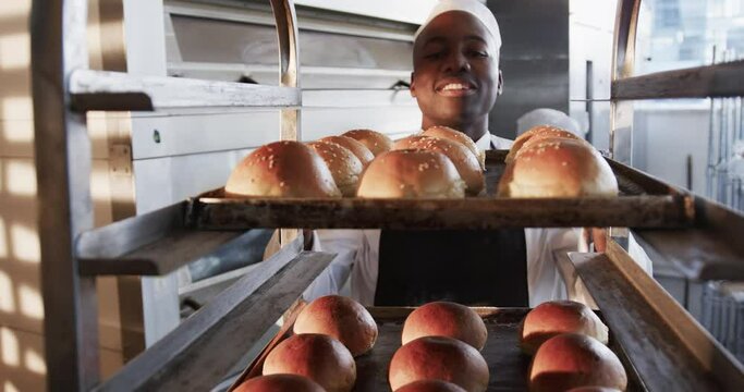 Happy african american male baker in bakery kitchen, holding baking sheet with rolls in slow motion