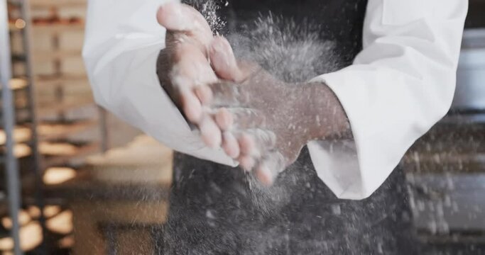 African american male baker working in bakery kitchen, brushing flour from hands in slow motion