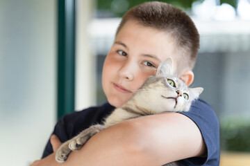 close-up of teenager looking at camera. Boy holding and gently hugging his tabby cat outdoors near...