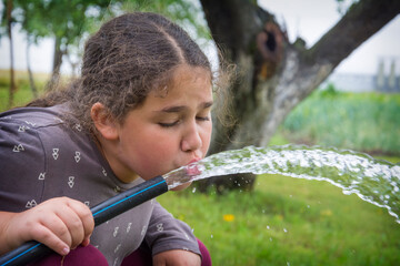 In summer, a girl drinks water from a hose in the garden.