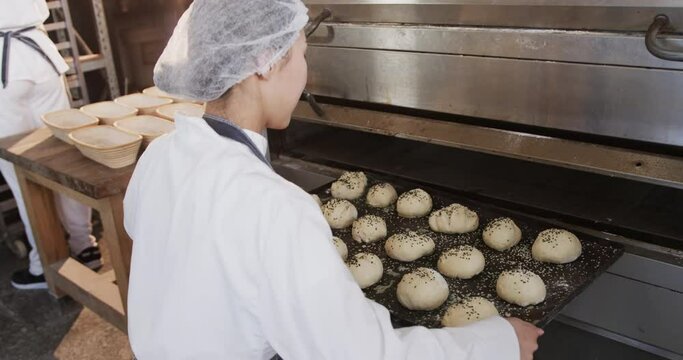 Asian female baker working in bakery kitchen, putting rolls on trays in oven, slow motion