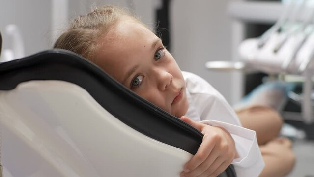 Close-up portrait of afraid little girl lying in dental chair sadness looking at camera unwilling to get dental treatment and turning her head away, slow motion. Concept of children teeth treatment