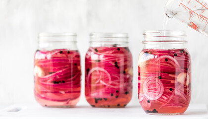 Preparing jars of pickled red onions with vinegar being poured into one.
