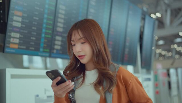 Happy asian woman traveller checking flight schedule departures board in airport terminal hall in front of check in counters. Tourist journey trip concept