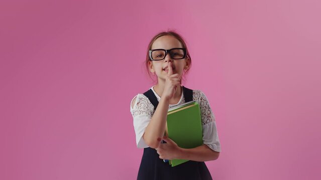 Portrait of a smart little girl kid wearing glasses presses index finger to lips makes silence hush gesture shh sign do not tells gossip secret, posing over colourful background wall in studio