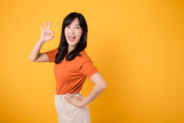 Radiating positivity, cheerful Asian woman 30s wearing orange shirt shows okay hand gesture against yellow background, symbolizing approval and assurance. Positivity, agreement, confidence concept.