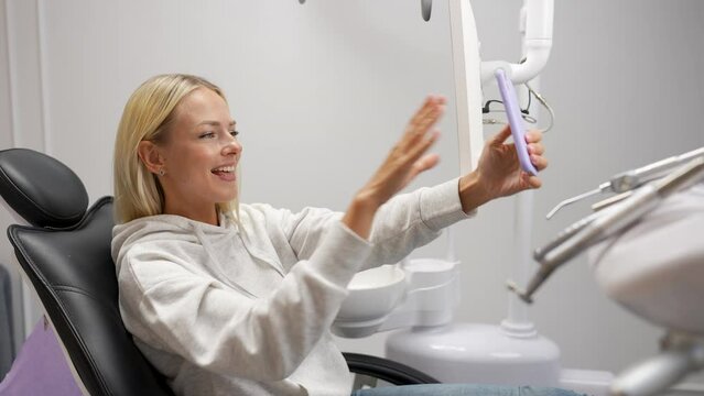 Happy blonde young woman talking on video call via mobile phone sitting in dental chair while waiting for dentist in modern dentistry clinic. Side view of female patient taking selfie video on phone.