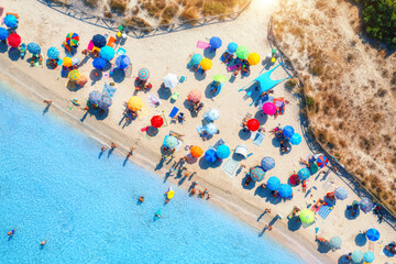 Aerial view of colorful umbrellas on sandy beach, swimming people in blue sea at sunrise in summer. Sardinia, Italy. Tropical landscape with clear azure water. Travel and vacation. Top drone view