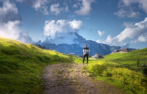 Girl on the rural road with yellow flowers and green grass in beautiful alpine mountain valley at sunset in summer. Young woman in old alpine village, sky with clouds. Travel and Hiking. Slovenia