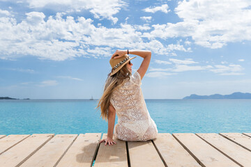 Smiling blonde woman with hat sitting in front of the turquoise sea in Mallorca