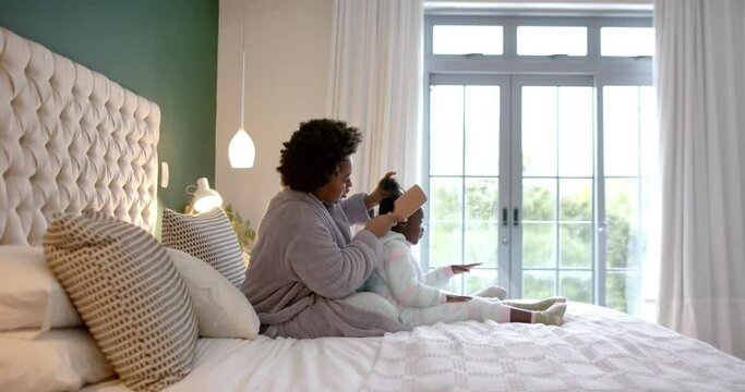 Happy african american mother combing daughter hair sitting on bed in bedroom, slow motion