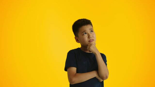 Young boy thinking stroking his chin with a hand on a yellow background. Concept of searching for interesting and useful activities for children