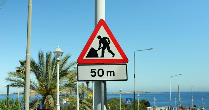 Road work sign with attached fifty meters distance plate mounted at lamp mast. Traffic sign warning about roadworks or construction ahead with sea and exotic palm tree in background in warm country.