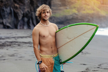 Handsome fit young blond man with mock up surfboard waits for wave to surf spot at sea ocean beach with black sand and looks at camera. Concept of sport, fitness, freedom, happiness, new modern life