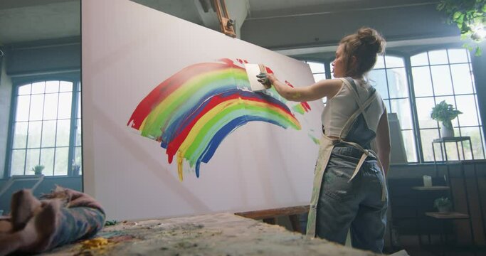 Millennial girl artist paints picture in loft studio using spatula to apply paint to canvas in daylight. Rainbow in the painting, cinematic. Painter concept, happy young multi ethnic artist, LGBTQ