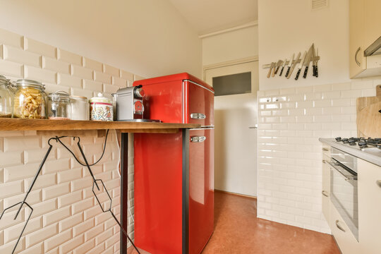 a small kitchen with red refrigerator and white tiles on the wall behind it is a wooden shelf that holds various uts