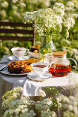 Fancy white porcelain set for herbal tea or coffee and homemade pie on wooden table in the garden. Summer outdoor party arrangement, romantic date. White flowers on background and in a glass vase