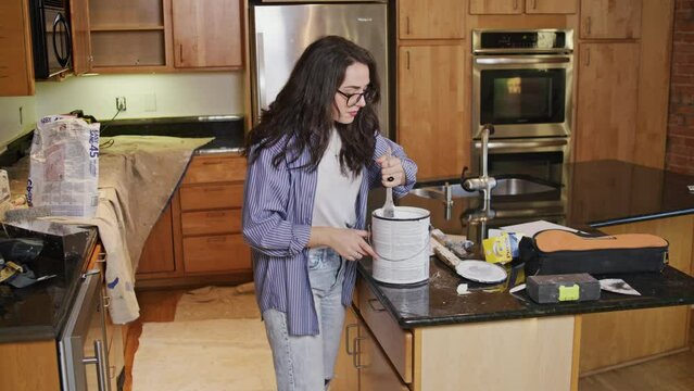 Young woman with glasses and a blue shirt prepares to paint her walls by stirring up a can of primer