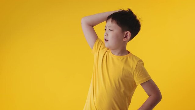 Portrait of a asian kid boy gesturing with raised hand touches his head, being buffled and confused, posing isolated over yellow color background wall in studio