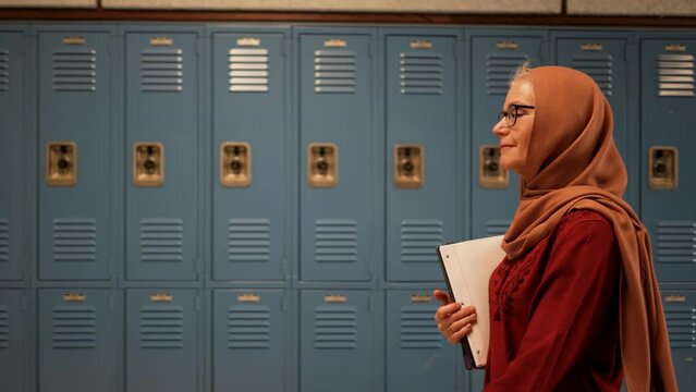 Slow motion side view of happy smiling muslim teacher wearing headscarf walking down a school hallway holding books with lockers in the background.