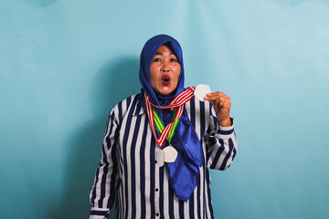 An excited middle-aged Asian woman in a blue hijab and a striped shirt is showcasing a medal while being isolated on a blue background