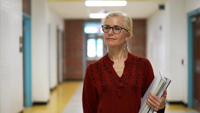 Closeup slow motion of pretty mature woman teacher holding books walking down an empty school hallway.