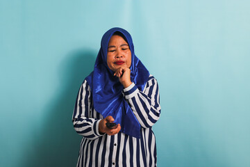 A bored middle-aged Asian woman in a blue hijab and a striped shirt is watching a boring TV show and switching channels with a remote control. She is isolated on a blue background.