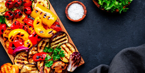 Grilled vegetables on rustic wooden cutting board,: colorful paprika, zucchini, eggplant, mushrooms, tomatoes and red onions with yogurt dressing, black stone table background, top view