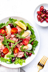 Nicoise salad with canned tuna, cherry tomatoes, boiled eggs, green beans and red olives on plate, white table background, top view